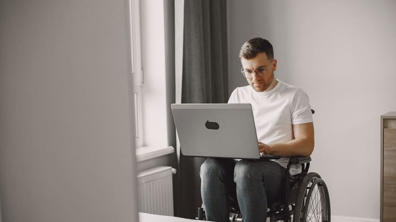 A man in a wheelchair typing on a laptop in a bright room