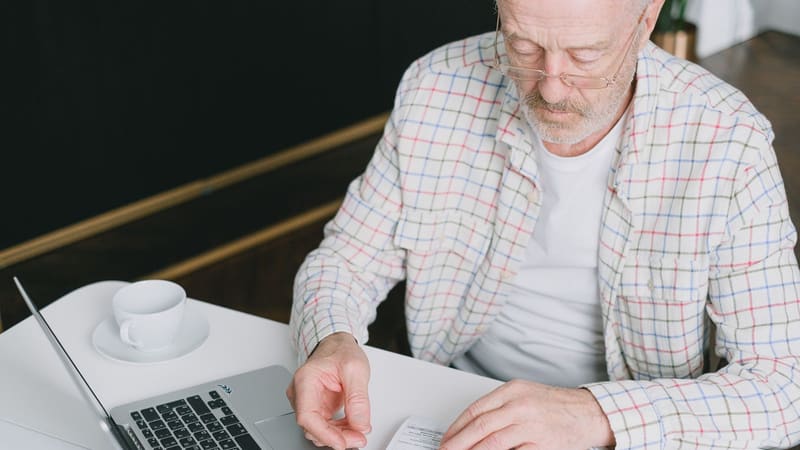 An older man in glasses reading a letter beside a laptop