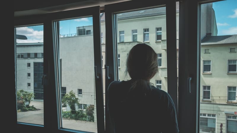 A woman standing at a window in her apartment, looking out