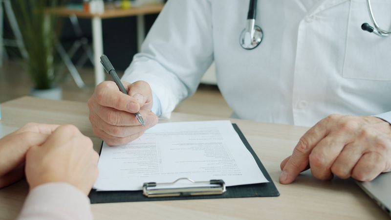 A doctor filling in a form on a clipboard while a patient sits across the desk