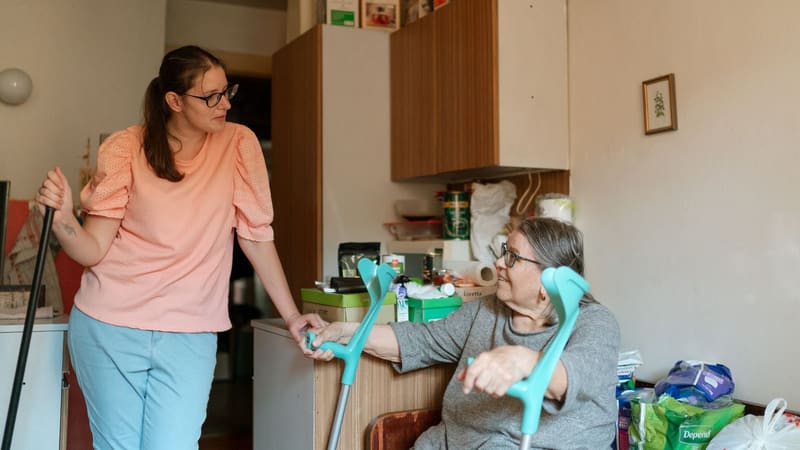 A caregiver helping an older woman with a crutch in her living room